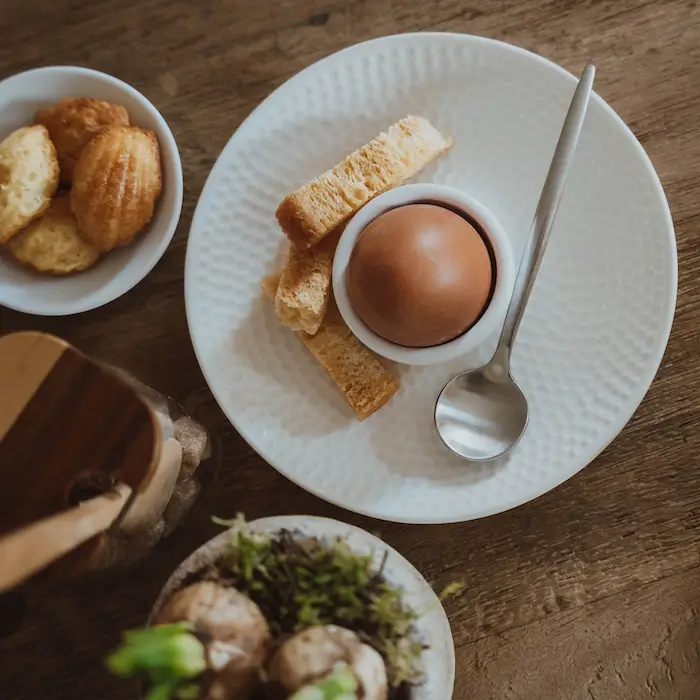 Ein weich gekochtes Ei in einem weißen Eierbecher auf einem weißen Teller, serviert mit drei Streifen geröstetem 
                            Brot und einem silbernen Löffel. Daneben steht eine kleine Schale mit Madeleines, eine Holzgewürzmühle und ein Topf 
                            mit Moos und Zwiebelpflanzen auf einem rustikalen Holztisch.