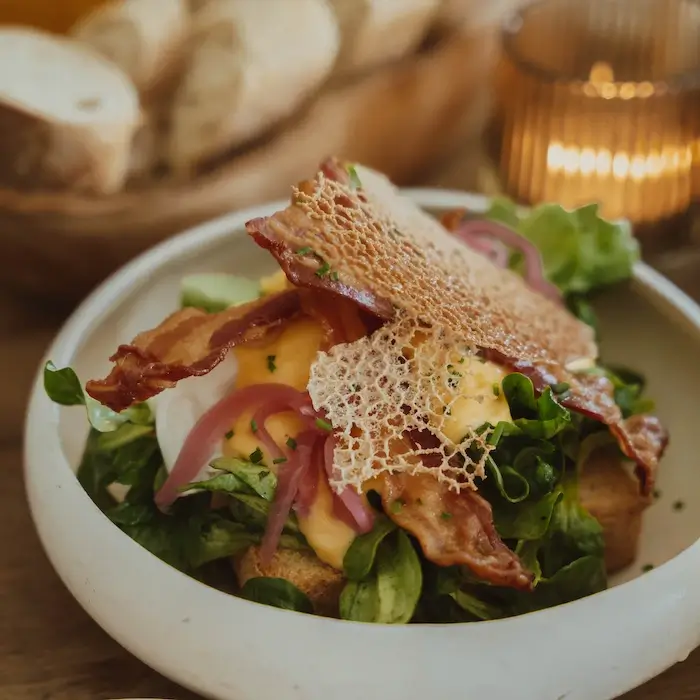 Ein kunstvoll angerichtetes Gericht in einer weißen Schüssel. Es besteht aus knusprigem Bacon, 
                            pochiertem Ei mit cremiger Sauce, frischem grünem Salat, eingelegten roten Zwiebeln und einem knusprigen 
                            Käsechip auf geröstetem Brot. Im Hintergrund sind ein Brotkorb und eine brennende Kerze zu sehen.
