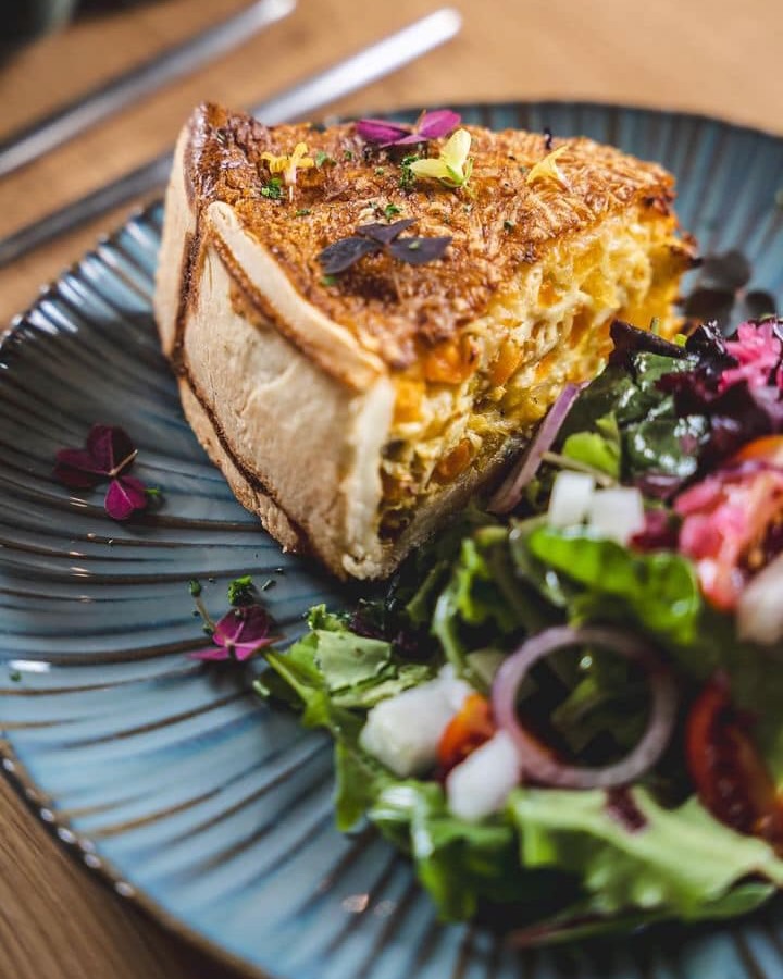 Ein Stück goldbraun gebackene Quiche auf einem blauen Teller, begleitet von einem frischen Salat 
                            aus Blattsalat, Tomaten, Zwiebeln und essbaren Blüten.