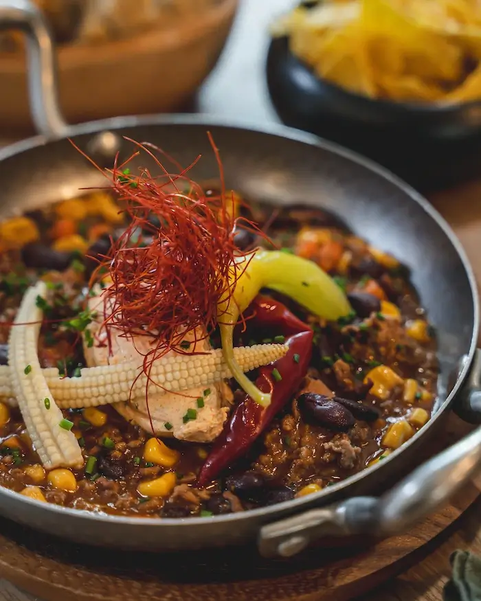 Ein würziges Gericht in einer schwarzen Pfanne, bestehend aus einer herzhaften Mischung aus Fleisch, 
                            Bohnen, Mais und Gewürzen. Garniert mit Babymais, roten und grünen Chilis, dünnen Chilifäden und einem Stück 
                            Hähnchen. Im Hintergrund sind ein Brotkorb und eine Schale mit knusprigen Tortillachips zu sehen.